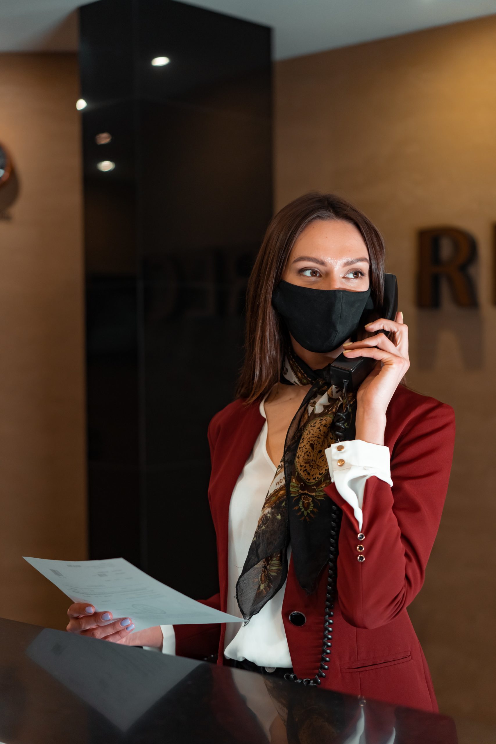 woman on the phone wearing a white shirt and red blazer wearing a mask, holding a piece of paper in front of a desk